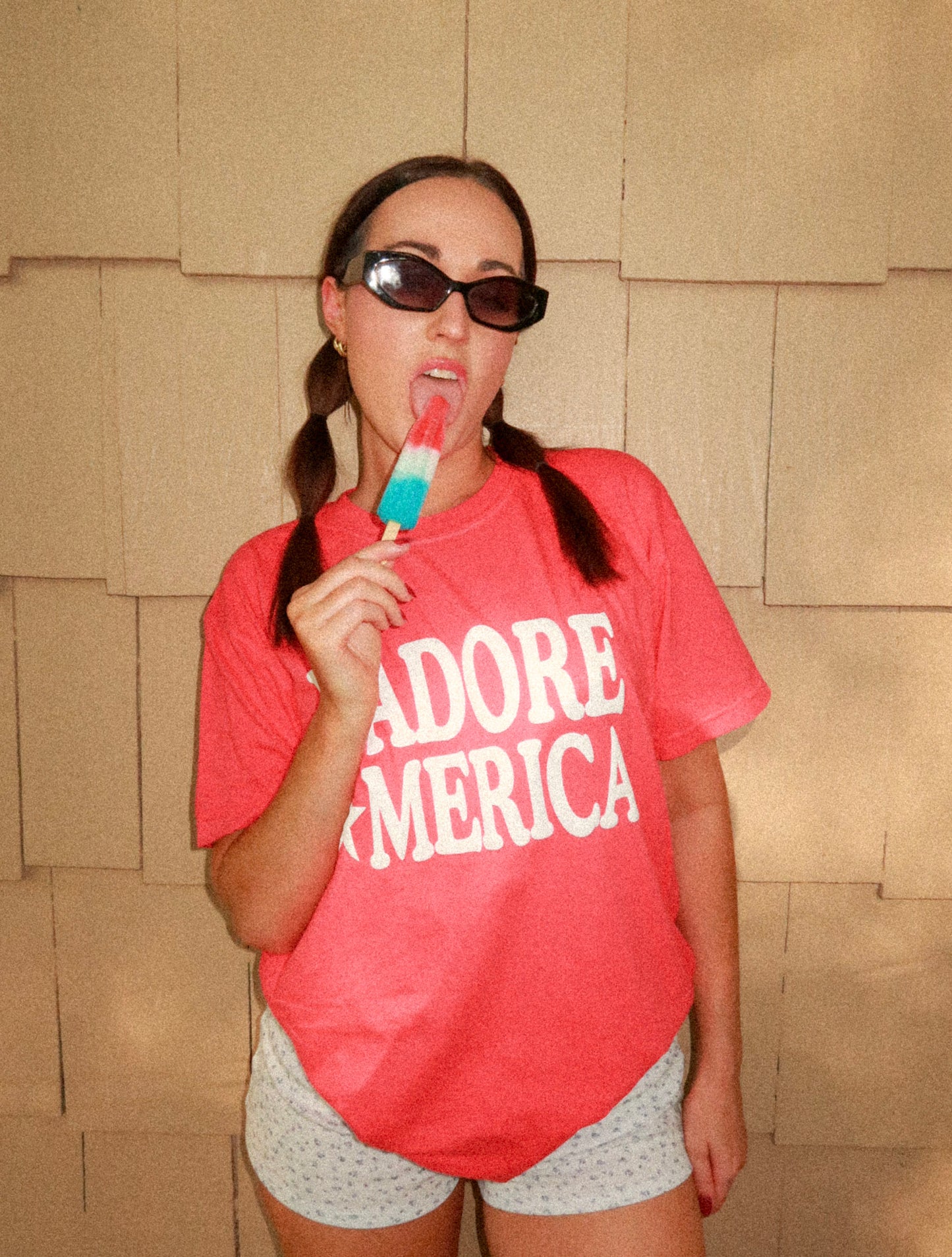 Model holding popsicle with full view of J’ADORE *MERICA shirt against blank wall